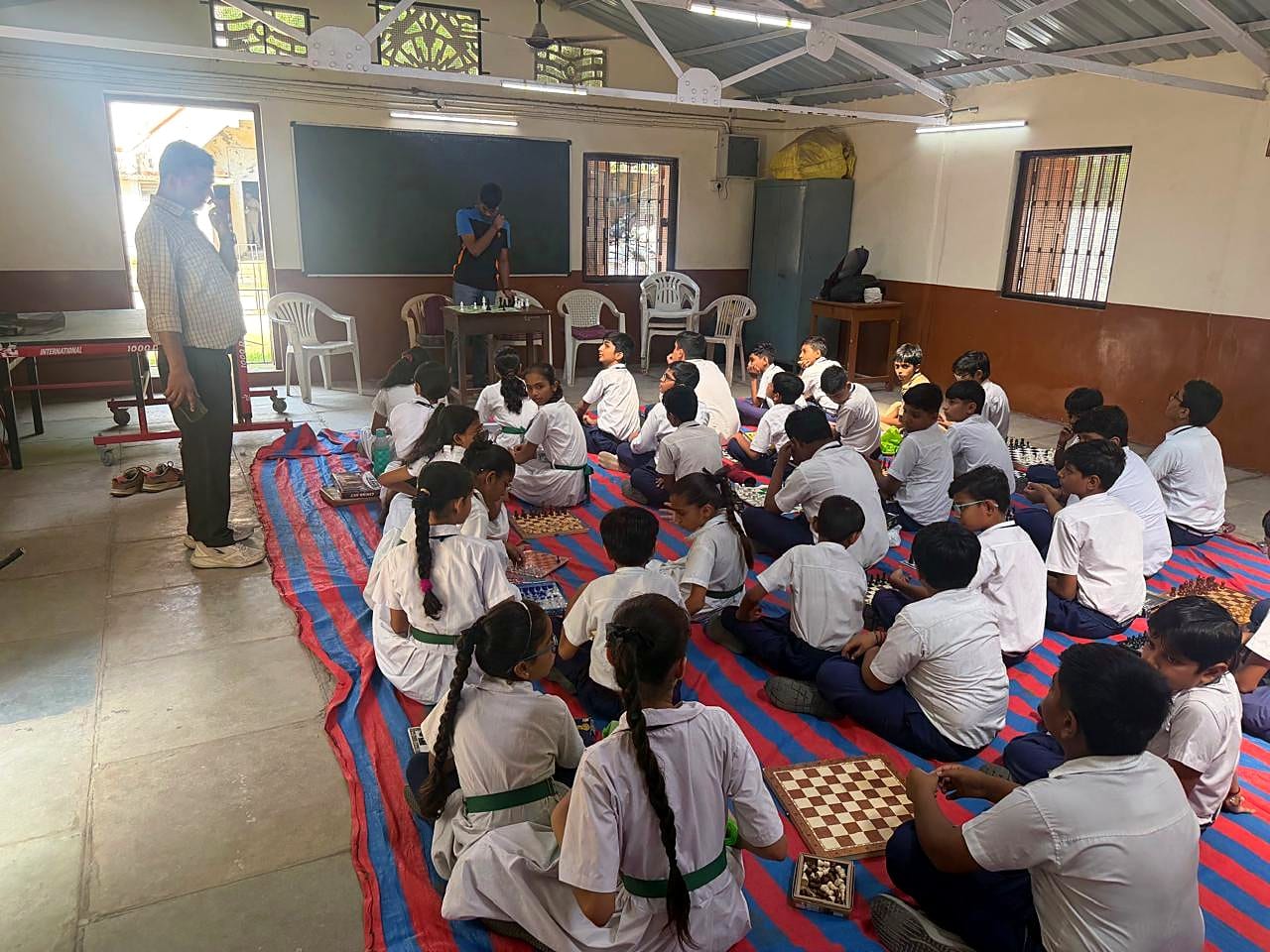 School students seated on a striped mat with chessboards, facing the teaching area.