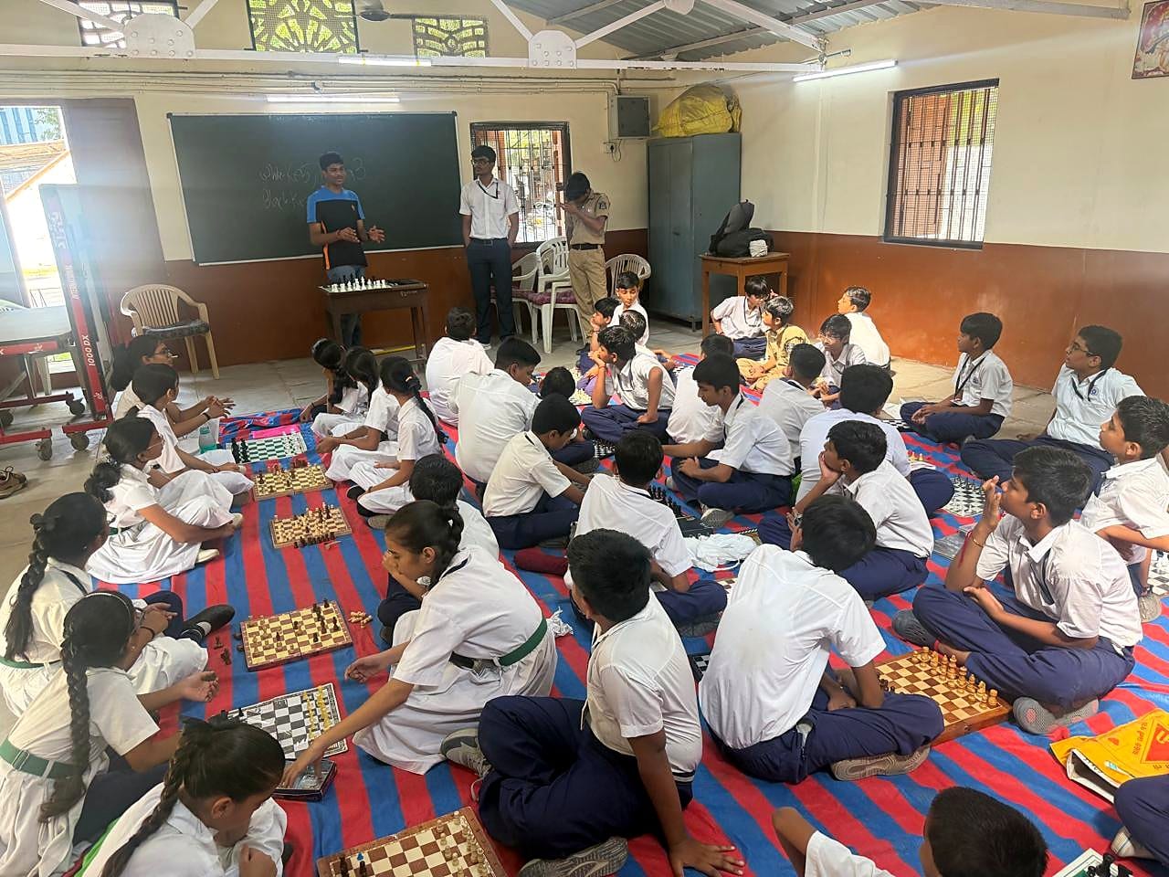 Mukund stands at a table with chess pieces in front of a blackboard while students sit on a striped mat facing him.